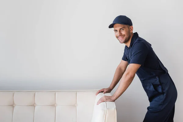 Счастливый рабочий, стоящий рядом с белым диваном и улыбающийся камере — стоковое фото