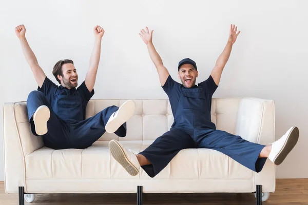 Взволнованные грузчики сидят на белом диване с поднятыми руками и ногами — стоковое фото