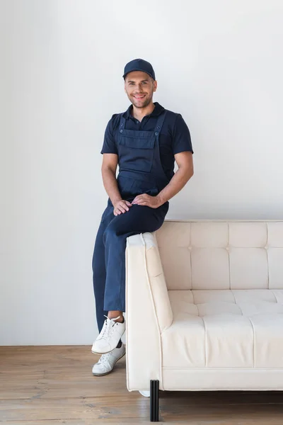 Happy worker in overalls smiling at camera on white couch — Stock Photo