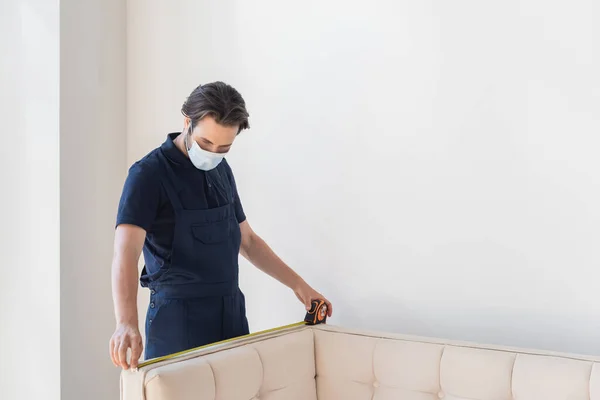 Worker in medical mask measuring couch near white wall — Stock Photo