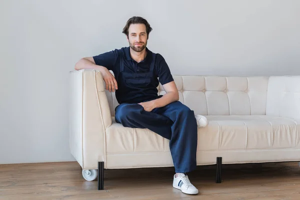 Вид в полный рост рабочего в комбинезоне, сидящего на белом диване — стоковое фото