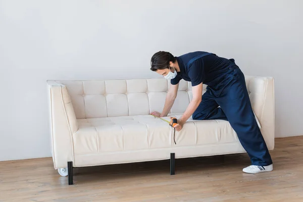 Bricoleur en masque de sécurité mesurant canapé près du mur blanc dans le salon — Photo de stock