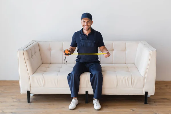 Full length view of smiling handyman sitting on white couch with measuring tape — Stock Photo