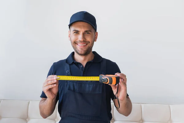 Cheerful repairman in cap showing measuring tape while smiling at camera — Stock Photo