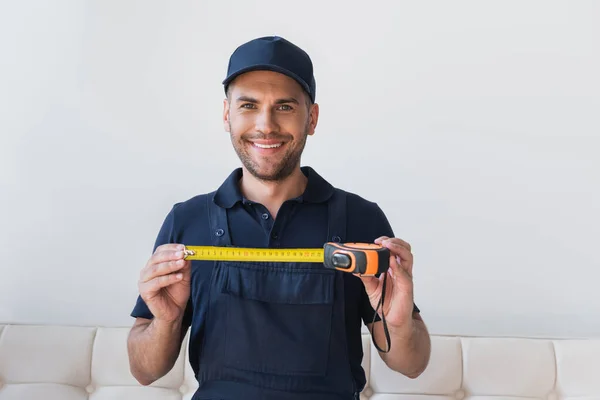 Réparateur joyeux dans le capuchon montrant ruban à mesurer tout en souriant à la caméra — Photo de stock