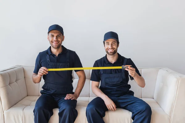 Cheerful movers sitting on sofa and holding measuring tape — Stock Photo