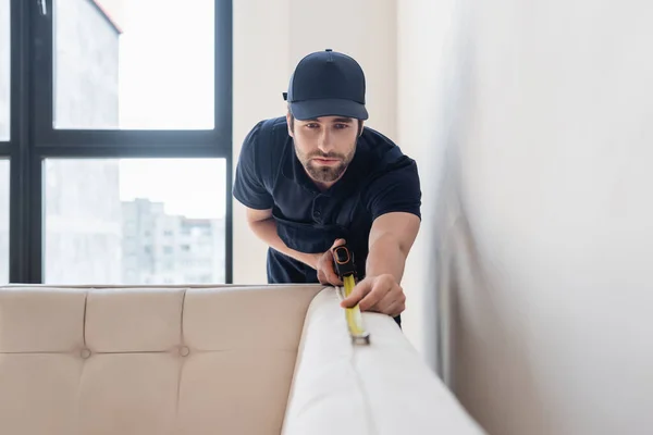 Young workman in cap measuring couch in modern apartment — Stock Photo