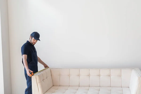 Young mover in cap and overalls measuring couch near white wall — Stock Photo