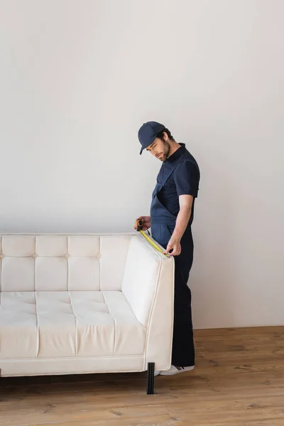 Ouvrier en uniforme mesurant canapé blanc dans le salon — Photo de stock