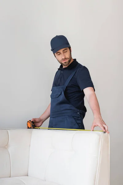 Foreman in cap and overalls with measuring tape near white couch — Stock Photo