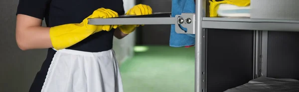 Cropped view of young chambermaid in apron holding handle of housekeeping cart, banner — Stock Photo