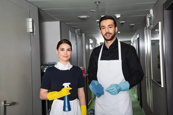 Cheerful housekeepers in rubber gloves looking at camera in corridor of hotel — Stock Photo