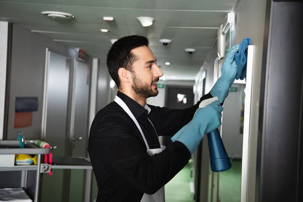 Side view of cheerful and bearded housekeeper in rubber gloves cleaning mirror in corridor of hotel — Stock Photo