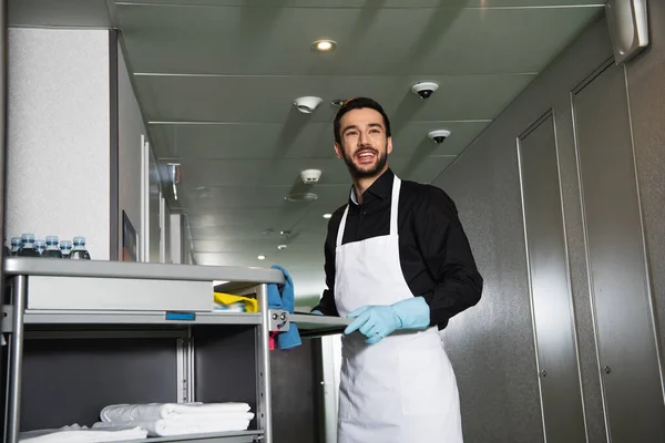 Cheerful and bearded housekeeper in rubber gloves standing near cart in corridor of hotel — Stock Photo