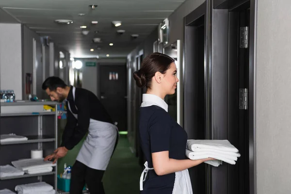 Vista lateral de empregada alegre em uniforme segurando lençóis limpos perto colega borrado perto de carrinho de limpeza — Fotografia de Stock