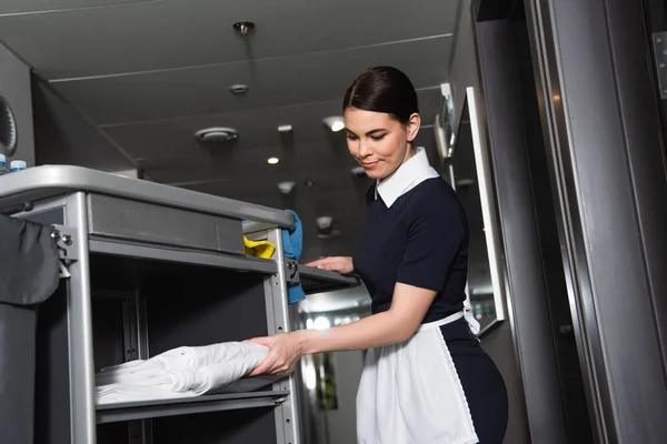 Brunette maid in uniform taking clean bed sheets from housekeeping cart — Stock Photo