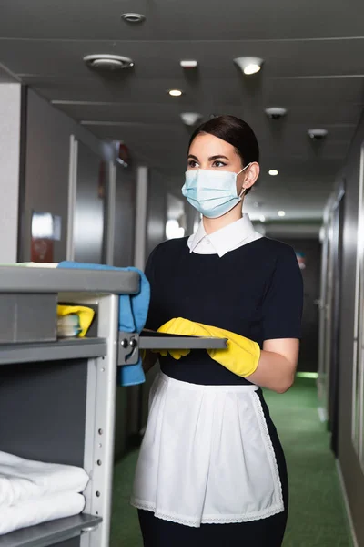 Giovane cameriera in maschera medica con maniglia del carrello delle pulizie — Foto stock