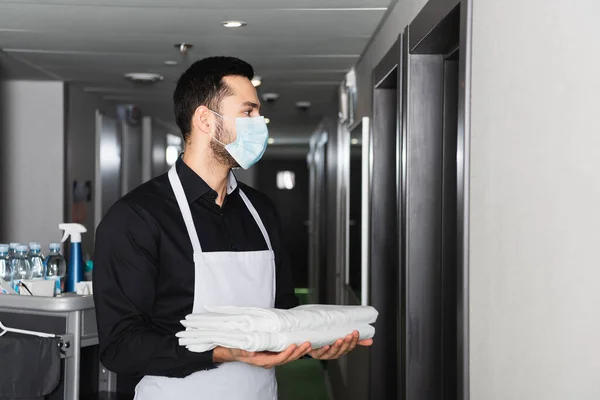 Housekeeper in medical mask holding clean bed sheets in corridor of hotel — Stock Photo