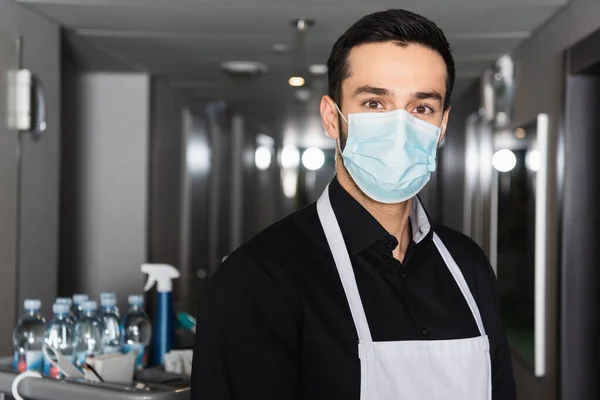 Housekeeper in apron and medical mask looking at camera in corridor of hotel — Stock Photo
