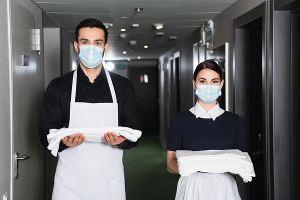 Haushälterinnen in Uniform und medizinischen Masken halten saubere Bettwäsche im Hotelflur — Stockfoto