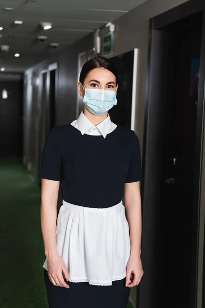 Junges Zimmermädchen in medizinischer Maske und Uniform blickt im Hotelflur in die Kamera — Stockfoto