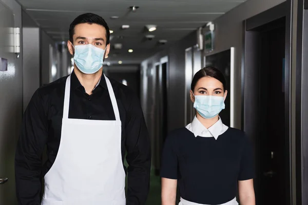 Young housekeepers in uniform and medical masks looking at camera in corridor of hotel — Stock Photo