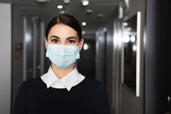 Jovem empregada doméstica em máscara médica e uniforme olhando para a câmera no corredor do hotel — Fotografia de Stock