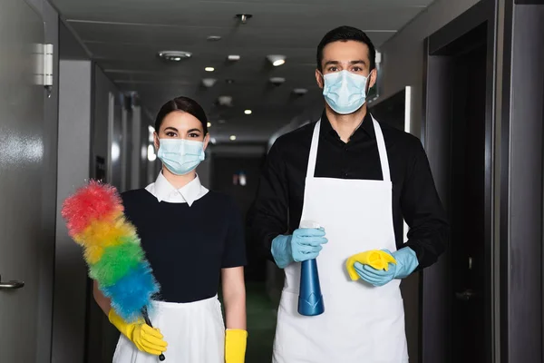 Empregadas domésticas em máscaras médicas e luvas de borracha segurando frasco de spray com pano e escova de poeira — Fotografia de Stock