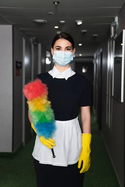 Empregada doméstica em máscara médica e luvas de borracha segurando escova de poeira no corredor do hotel — Fotografia de Stock