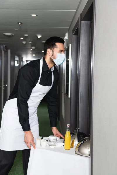 Waiter in medical mask and apron delivering tray with cloche and drinks in hotel — Stock Photo