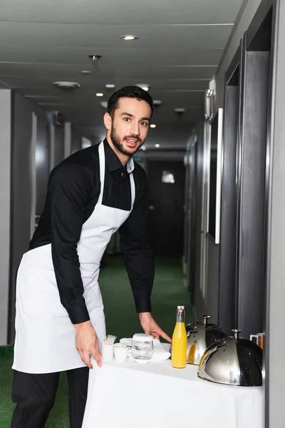 Serveur gai dans le masque médical et tablier plateau de livraison avec cloche et boissons — Photo de stock