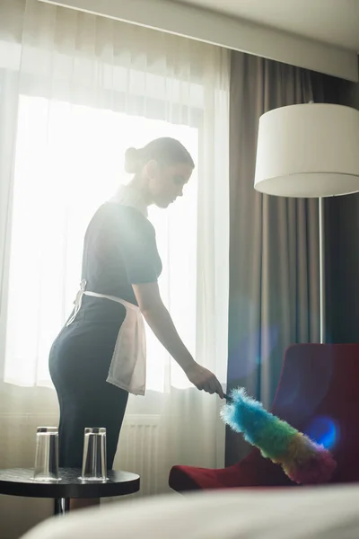 Jeune femme de ménage en uniforme de nettoyage fauteuil rouge avec brosse à poussière dans la chambre d'hôtel — Photo de stock