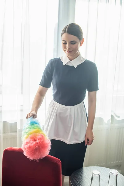 Camareira satisfeito em uniforme de limpeza poltrona vermelha com escova de poeira no quarto do hotel — Fotografia de Stock
