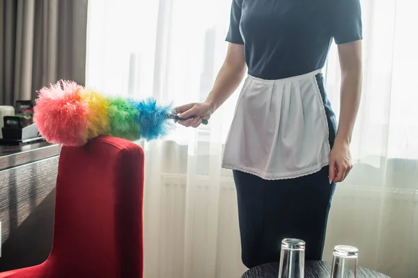 Vue recadrée de la femme de chambre en uniforme de nettoyage fauteuil rouge avec brosse à poussière à l'hôtel — Photo de stock