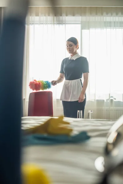 Femme de chambre en uniforme de nettoyage fauteuil rouge avec brosse à poussière — Photo de stock