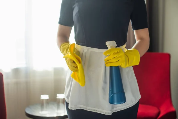 Vista recortada de la joven sirvienta en guantes de goma sosteniendo botella de spray y trapo - foto de stock