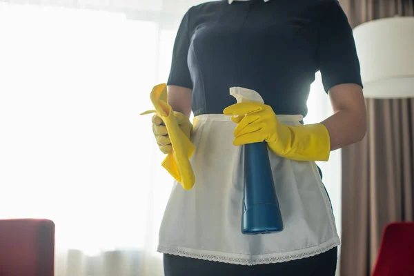 Visão parcial de jovem empregada doméstica em luvas de borracha segurando frasco de spray e pano — Fotografia de Stock