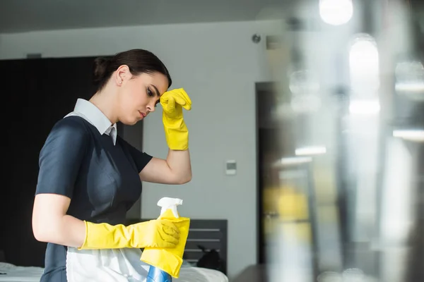 Tired chambermaid in rubber gloves holding spray bottle and wiping sweat while sitting on bed in hotel room — Stock Photo