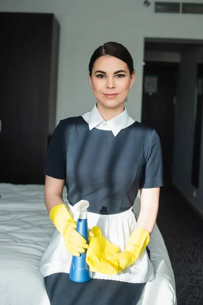 Gaie femme de chambre dans des gants en caoutchouc tenant bouteille de pulvérisation et assis sur le lit dans la chambre d'hôtel — Photo de stock