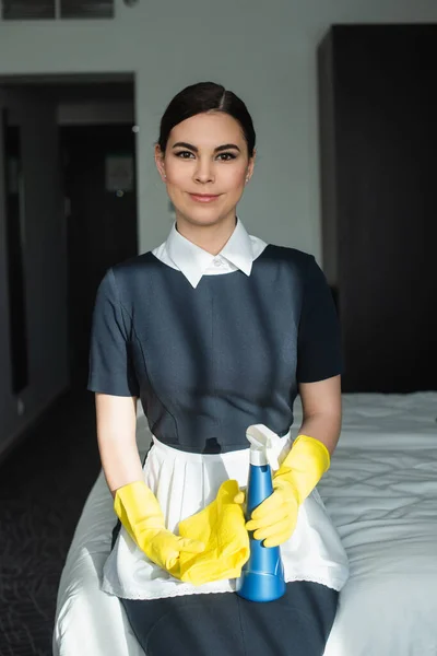 Sorrindo camareira em luvas de borracha segurando frasco de spray e sentado na cama no quarto do hotel — Fotografia de Stock