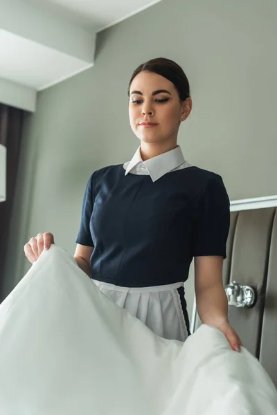 Joven camarera cambiando ropa de cama blanca en la habitación de hotel - foto de stock