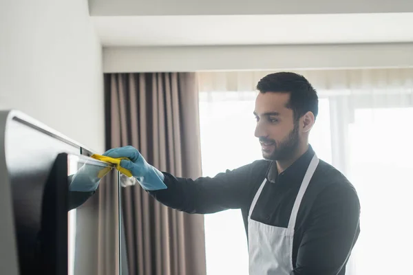 Bearded housekeeper in blue rubber glove cleaning tv display with rag — Stock Photo