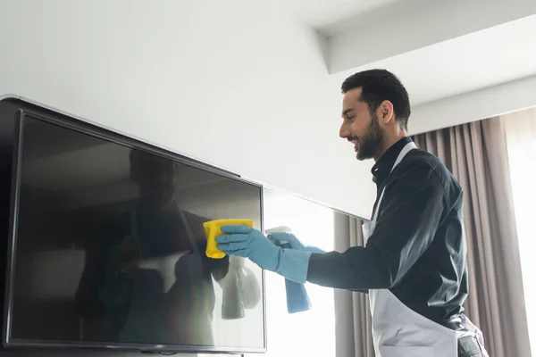 Vue latérale de la gouvernante gaie et barbu en gants de caoutchouc bleu nettoyage écran de télévision avec chiffon — Photo de stock