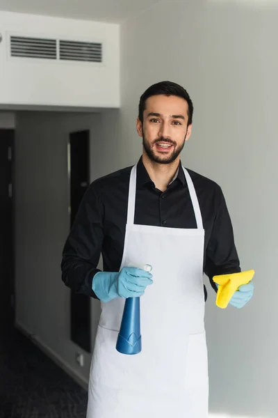Cheerful and bearded housekeeper in blue rubber gloves holding spray bottle and rag — Stock Photo