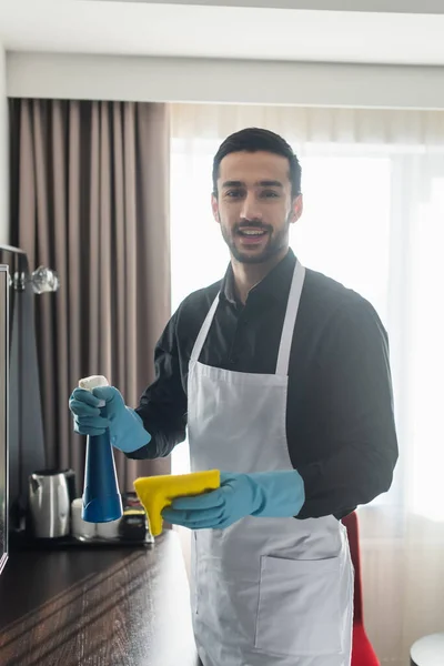 Positive and bearded housekeeper in blue rubber gloves holding spray bottle and rag — Stock Photo