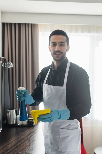 Governante positivo e barbuto in guanti di gomma blu con bottiglia spray e straccio — Foto stock