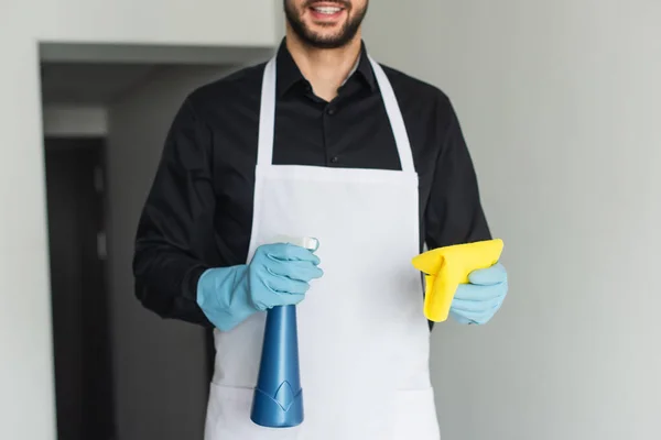 Cropped view of cheerful and bearded housekeeper in blue rubber gloves holding spray bottle and rag — Stock Photo