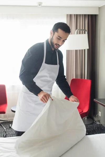 Sorridente e barbudo governanta mudando roupa de cama branca no quarto de hotel — Fotografia de Stock