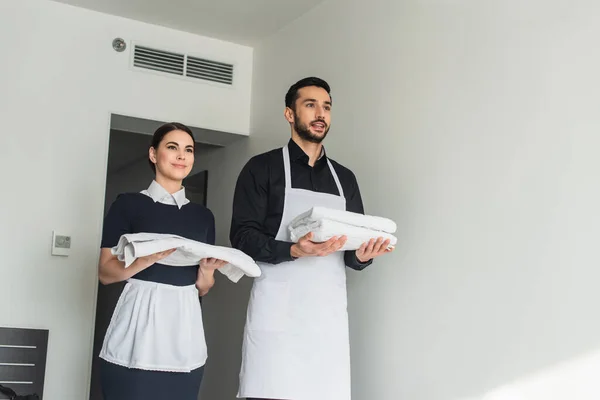 Alegre ama de llaves con sábanas blancas y toallas en la habitación de hotel - foto de stock