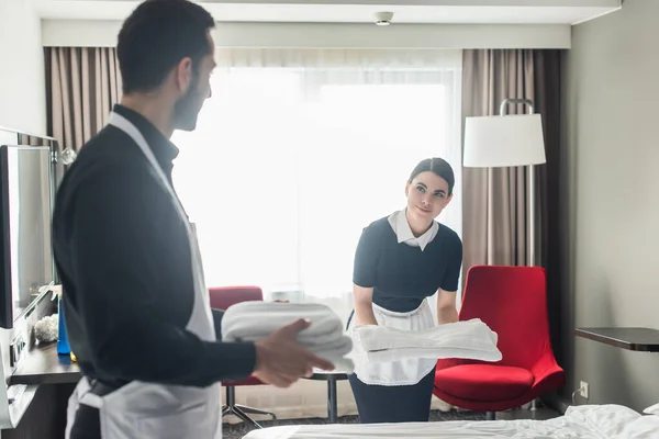 Young maid holding white bed sheets near colleague with clean towels on blurred foreground — Stock Photo